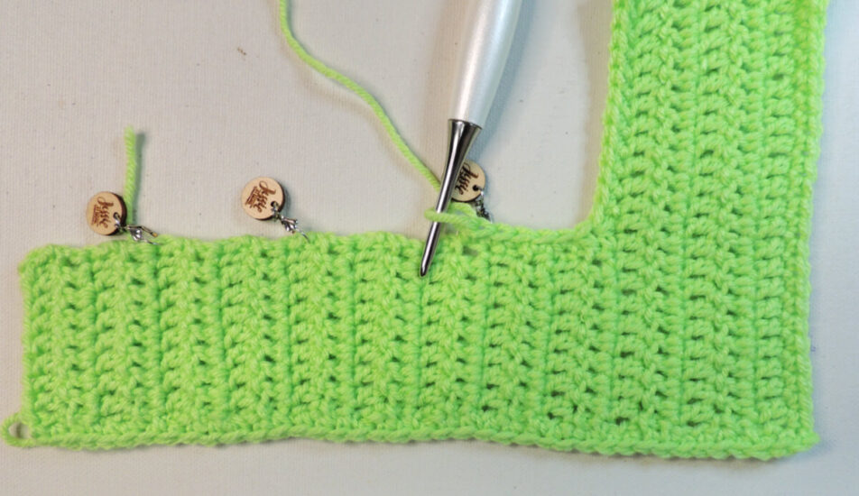 An "L" shape made of green crochet with the long edge at the bottom. On the top of the long edge we can see are 3 stitch markers and a crochet hook about a fourth of the way across making a border.