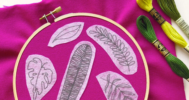 Fushia fabric stretched in a wood embroidery hoop with pieces of white water-soluble fabric stuck on it. There are leaf outline drawings on the white fabric. Next to the hoop are green and yellow skeins of embroidery floss.