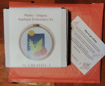 Small white box with label reading "plants and shapes applique embroidery kit" and "M Creative J". Under box is a notecard with "Way to get stitchy!" printed on the top, and more text underneath. Below that is a coral shipping padded envelope. All of that is on a wood table top.