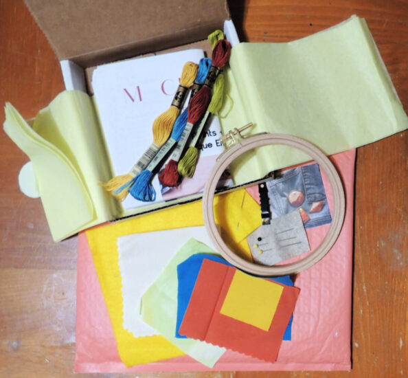 A coral shipping padded envelope on top of a wood table. On top of that is an open thing white box with yellow tissue paper lining opened up. On top of all of that are various pieces of fabric, embroidery floss, pins and needles, an embroidery hoop, a picture hanger, and instructions; all from the box.