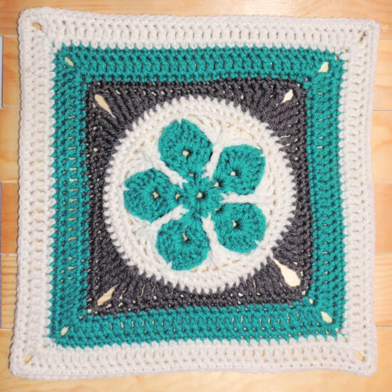 Crochet square with a teal 5 point flower in a white circle in the center, then a round of gray to make a square, followed by rounds in teal than white.