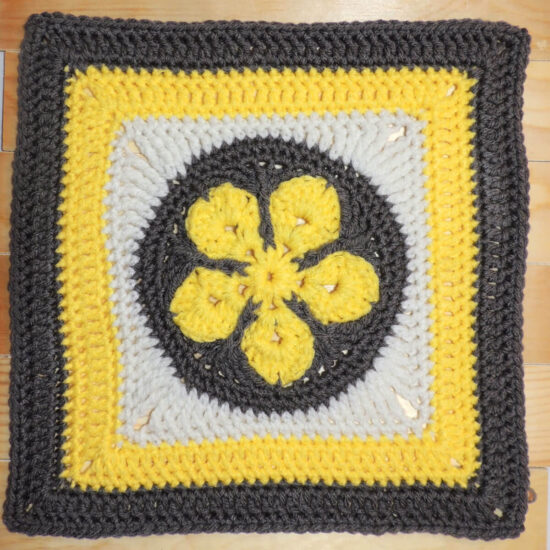 Crochet square with a yellow 5 point flower in a gray circle in the center, then a round of white to make a square, followed by rounds in yellow than gray.