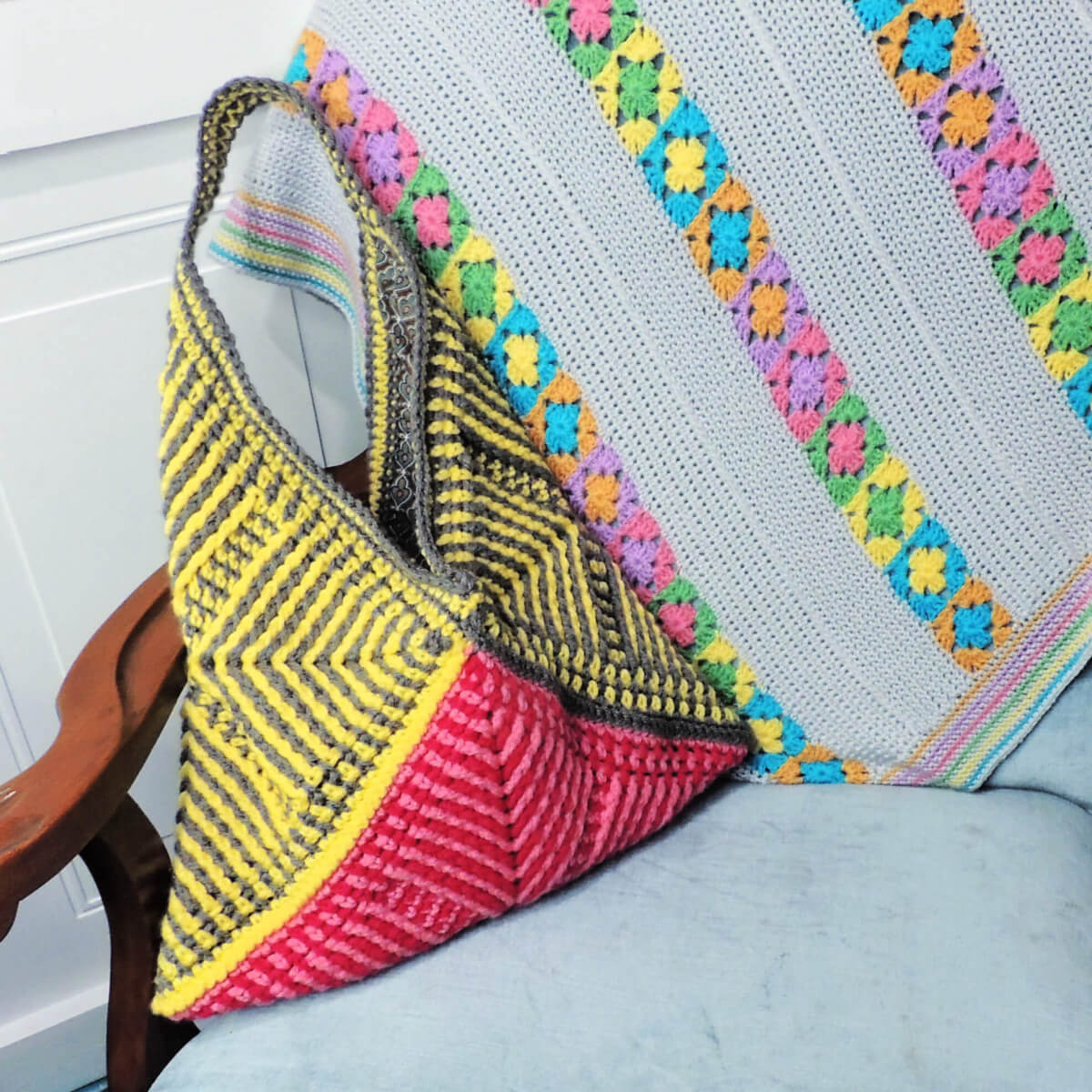 Purse made of three crochet squares folded diagonally. Each has angled stripes pointing in from corners made with post stitches. The bottom is two tone pink and the rest is gray and yellow. Purse leaning against the wood arm of a light blue chair.