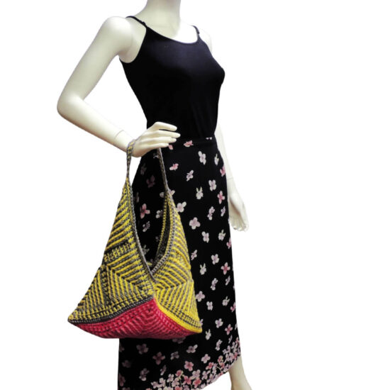 Side view of a white mannequin in a black tank top and black skirt with light flowers with a large purse hanging from its wrist. Purse is made of three crochet squares folded diagonally. Each has angled stripes pointing in from corners made with post stitches. The bottom is two tone pink and the rest is gray and yellow.