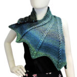 Front view of the top half of a white mannequin wearing a black tank top and a blue and green variegated knit buttoned cowl that is thin on one end then wraps around the neck and buttons to the other end which has flared out with 5 points. There are columns of raised large stitches from the short end to the tips. The cowl is buttoned on the mannequin's right shoulder.
