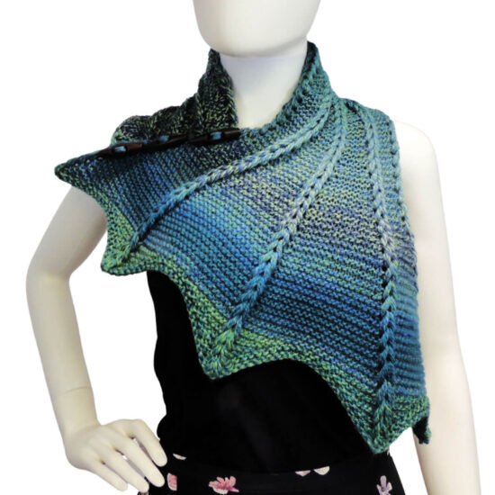 Front view of the top half of a white mannequin wearing a black tank top and a blue and green variegated knit buttoned cowl that is thin on one end then wraps around the neck and buttons to the other end which has flared out with 5 points. There are columns of raised large stitches from the short end to the tips. The cowl is buttoned on the mannequin's right shoulder.
