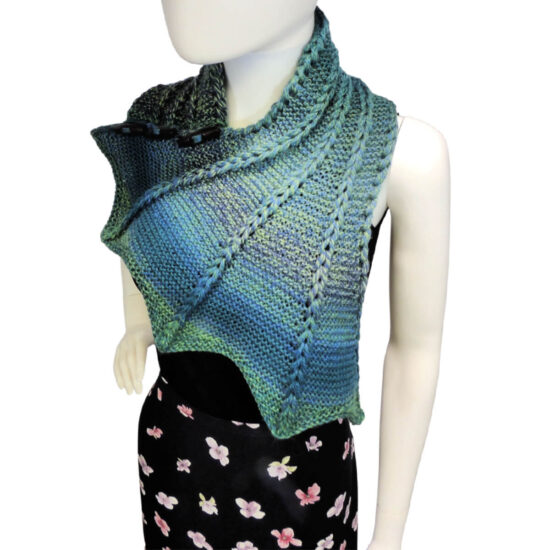 Raised front view of a white mannequin wearing a black tank top, black floral skirt and a blue and green variegated knit buttoned cowl that is thin on one end then wraps around the neck and buttons to the other end which has flared out with 5 points. There are columns of raised large stitches from the short end to the tips. The cowl is buttoned on the mannequin's right shoulder.
