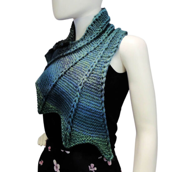 Three quarter front view of the top half of a white mannequin wearing a black tank top and a blue and green variegated knit buttoned cowl that is thin on one end then wraps around the neck and buttons to the other end which has flared out with 5 points. There are columns of raised large stitches from the short end to the tips. The cowl is buttoned on the side facing away from us.