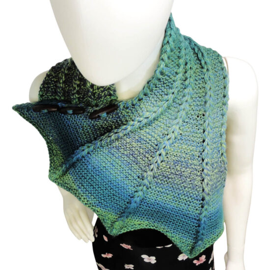 Up high front view of the top half of a white mannequin wearing a black tank top and a blue and green variegated knit buttoned cowl that is thin on one end then wraps around the neck and buttons to the other end which has flared out with 5 points. There are columns of raised large stitches from the short end to the tips. The cowl is buttoned on the mannequin's right shoulder.