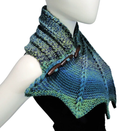 Side view with a bit of a tilt to the front of the top half of a white mannequin wearing a black tank top and a blue and green variegated knit buttoned cowl that is thin on one end then wraps around the neck and buttons to the other end which has flared out with 5 points. There are columns of raised large stitches from the short end to the tips. The cowl is buttoned on the side facing us.