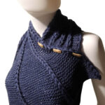 Three quarter front view of the top third of a white mannequin wearing a black tank top and a dark blue knit buttoned cowl that is thin on one end then wraps around the neck and buttons to the other end which has flared out with 5 points. There are columns of raised large stitches from the short end to the tips. The cowl is buttoned on the side facing us.