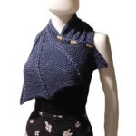 Three quarter front view of the top half of a white mannequin wearing a black tank top and a dark blue knit buttoned cowl that is thin on one end then wraps around the neck and buttons to the other end which has flared out with 5 points. There are columns of raised large stitches from the short end to the tips. The cowl is buttoned on the side facing us.