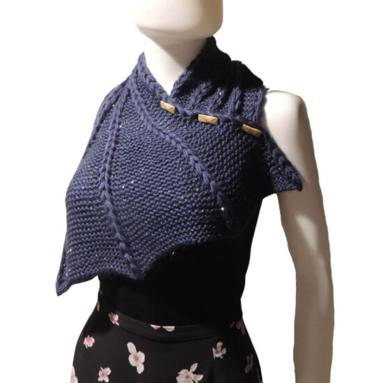 Three quarter front view of the top half of a white mannequin wearing a black tank top and a dark blue knit buttoned cowl that is thin on one end then wraps around the neck and buttons to the other end which has flared out with 5 points. There are columns of raised large stitches from the short end to the tips. The cowl is buttoned on the side facing us.