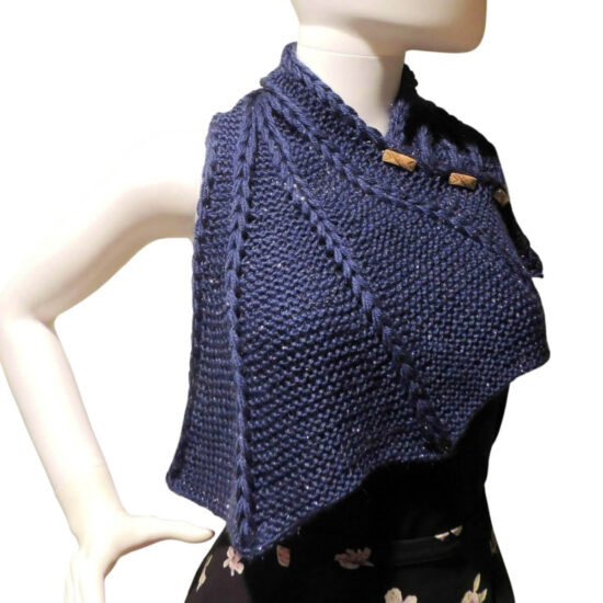 Front view of the top half of a white mannequin wearing a black tank top and a dark blue knit buttoned cowl that is thin on one end then wraps around the neck and buttons to the other end which has flared out with 5 points. There are columns of raised large stitches from the short end to the tips. The cowl is buttoned on the mannequin's left shoulder.