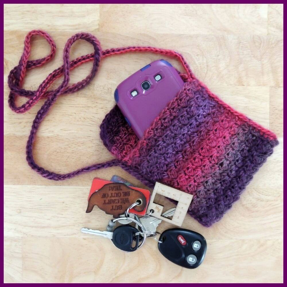 A small rectangular crochet bag with long cord strap, all in dark pink and purple color changing yarn, laying on a light wood table. In the bag is a purple phone, and next to the bag is a set of keys with keychains.