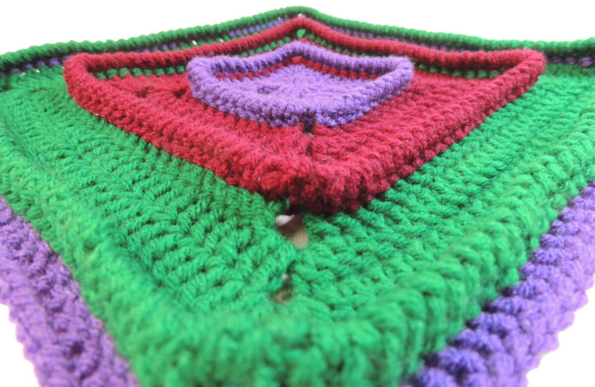 Close up angled view of a crochet square that starts with purple on the inside, then has a section of burgundy that is attached below the last round of the purple, making the last round stand up, then a section of green is attached in the same manner to the burgundy, and finally a thin section of purple attached in same manner.