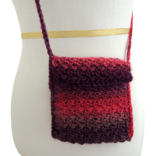 Close up of side of a white dress form with a small rectangular crochet bag with a cross body strap, all in dark pink and purple color changing yarn.