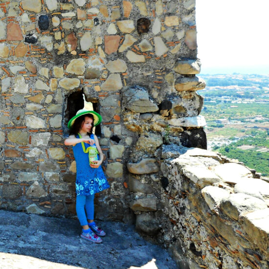 Square photo of a chile next to a wall on a castle, looking over a village. The child is wearing all blue and a yellow and green sun hat and is opening a water bottle that's in a lime green crochet mesh bottle holder with a cross body strap.