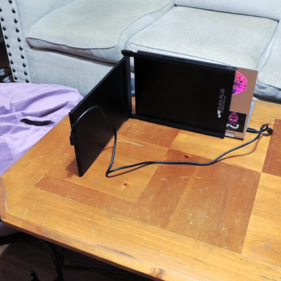 A slightly raised and back view of a laptop with a second screen on a table. The second screen is attached to the back of the laptop and folded to the side so that the front of the laptop is at the front of the table facing a couch and the second screen is at the side of the table facing a bean bag. A cord attaches the two.