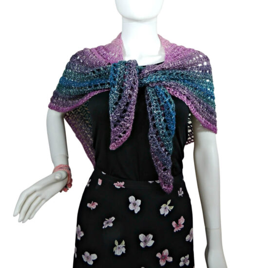 A white mannequin in a black tank top, black skirt and a triangular crochet shawl that changes colors through pink, purple, green, and blue. Shawl alternates between rows of solid stitches and rows of net or eyehole stitches. The point of the shawl is in back and the side points are pulled over the shoulders and tied in the front.