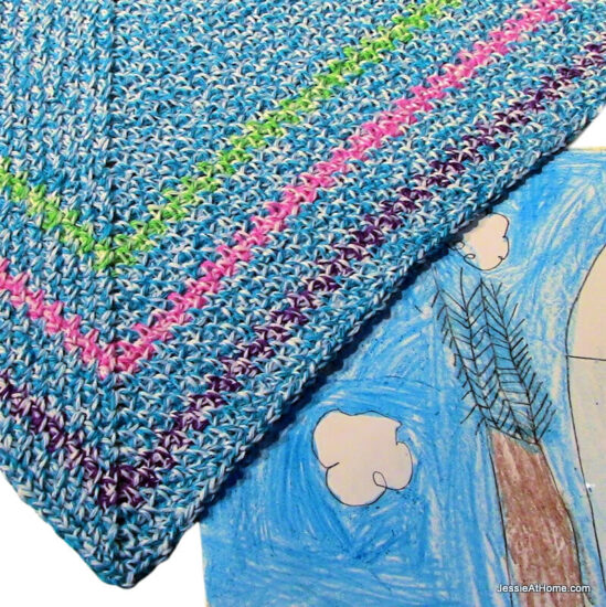 Close up overhead view of a crochet right angle 45/45/90 triangle. The triangle is light blue with 3 stripes close to the edge in green, pink, and purple. Under it is a bright children's painting of an outdoor winter scene in blue, white, and brown.