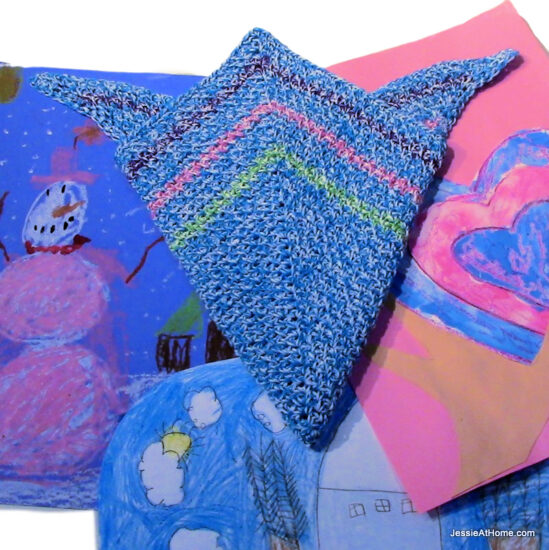 A crochet right angle 45/45/90 triangle bandana that has been folded in thirds so there is a point at the center of the long sides and all 3 triangle points are at the top. The bandanna is light blue with 3 stripes close to the edge in green, pink, and purple. Under it are 3 bright children's paintings, one of a pink and white snowman on blue paper, one of a pink and blue heart on pink paper, and the third is an outdoor winter scene in blue, white, and brown.