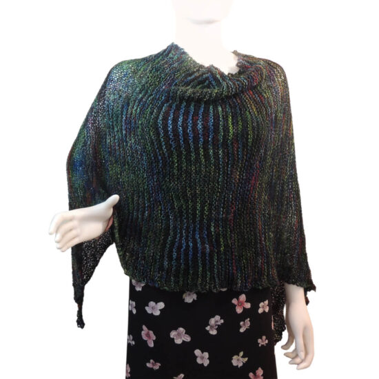 White mannequin wearing a black tank and skirt and a knit poncho in black with bits of dark red, blue, and green. The poncho is waist length and straight across the front with a point hanging off the right arm which is bent at the elbow.