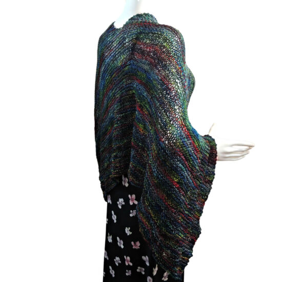 Side view of a white mannequin wearing a black tank and skirt and a knit poncho in black with bits of dark red, blue, and green. The poncho is waist length and straight across the front with a point hanging off one arm.