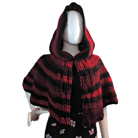 Front view of a white mannequin in a black skirt and a hooded waist length crocheted cape. The cape has cables down the front opening and horizontal stripes of black and red.