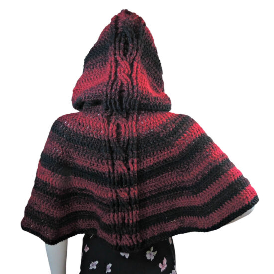 Back view of a white mannequin in a black skirt and a hooded waist length crocheted cape. The hood and cape have cables down the center back and horizontal stripes of black and red.