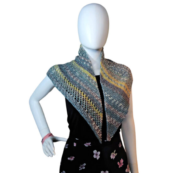 White mannequin wearing a black tank and skirt and a small triangular knit shawl. The shawl is knit in tweed yarn with long stripes of blue-gray and short stripes of yellow. The knitted fabric alternates between sections of garter stitch and a mesh stitch. The center point of the shawl is in back and the two sides drape over the shoulders.