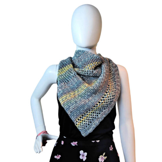 White mannequin wearing a black tank and skirt and a small triangular knit shawl. The shawl is knit in tweed yarn with long stripes of blue-gray and short stripes of yellow. The knitted fabric alternates between sections of garter stitch and a mesh stitch. The shawl point is in front and the sides are pulled behind the neck.