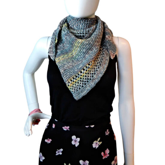 White mannequin wearing a black tank and skirt and a small triangular knit shawl. The shawl is knit in tweed yarn with long stripes of blue-gray and short stripes of yellow. The knitted fabric alternates between sections of garter stitch and a mesh stitch. The shawl point is in front and the sides are wrapped around the neck and hang short on either side of the front.