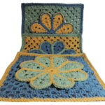 Image of 3 crochet granny squares, each with a single crochet border and an overlayed large flower with 8 petals each outlined in slip stitches. One is laying flat, another is partially standing behind the first and a third is standing behind the second. They are each in combinations of yellow, blue and green. The photo is taken at an angle so all can be seen.