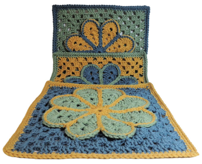 Image of 3 crochet granny squares, each with a single crochet border and an overlayed large flower with 8 petals each outlined in slip stitches. One is laying flat, another is partially standing behind the first and a third is standing behind the second. They are each in combinations of yellow, blue and green. The photo is taken at an angle so all can be seen.