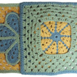 A crochet granny square with a single crochet border, overlayed on the square is a large flower with 8 petals each outlined in slip stitches. The square is yellow with a green border and the flower is blue with a green outline. Laying half way over it is a second square that's upside down and has the colors in a different order.