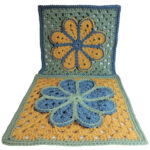 Image of 2 crochet granny squares, each with a single crochet border and an overlayed large flower with 8 petals each outlined in slip stitches. One is laying flat and the other is standing up behind the first. They are each in combinations of yellow, blue and green. The photo is taken at an angle so both can be seen.