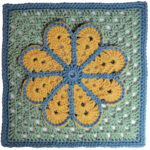 A crochet granny square with a single crochet border, overlayed on the square is a large flower with 8 petals each outlined in slip stitches. The square is green with a blue border and the flower is yellow with a blue outline.