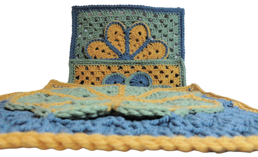 Image of 3 crochet granny squares, each with a single crochet border and an overlayed large flower with 8 petals each outlined in slip stitches. One is laying flat and the photo is taken across that one with 2 more behind each. They are each in combinations of yellow, blue and green.