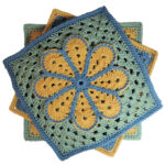 A crochet granny square with a single crochet border, overlayed on the square is a large flower with 8 petals each outlined in slip stitches. The square is yellow with a green border and the flower is blue with a green outline.