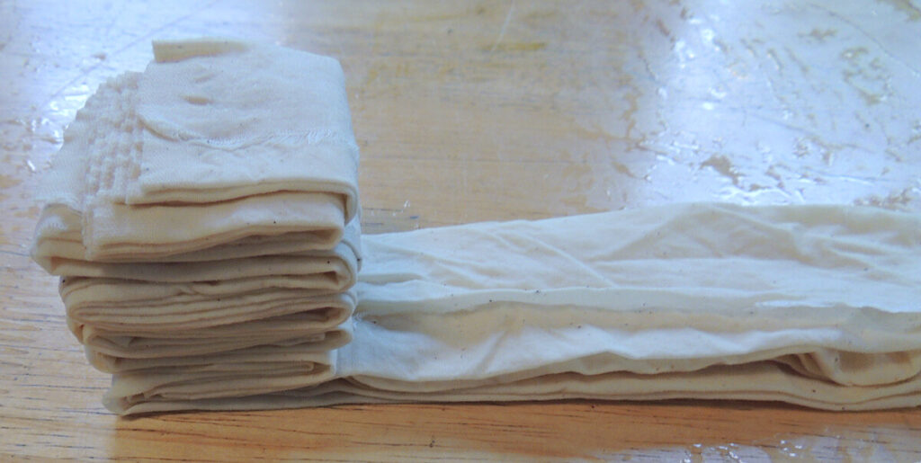 A piece of muslin fabric accordion folded two ways, sitting on a light wood table.