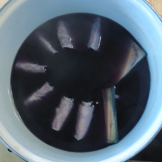 Overhead view of a white pot with blue dye inside. In the dye is accordion folded fabric, and we can see just the bumps of the folds in a circle above the dye.