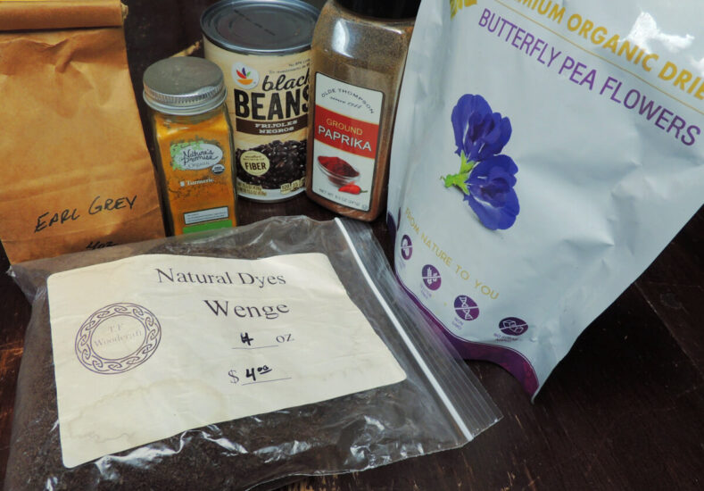 Items sitting on a dark table: small brown bag labeled "Earl Gray Tea", bottle of Turmeric, can of black beans, paprika, bag of dried butterfly pea flowers, bag of dark sawdust labeled "Natural Dye Wedge"