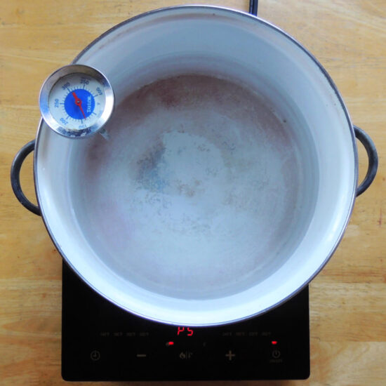 Overhead view of a large white pot on a black hotplate on a light wood table. A candy thermometer is attached to the pot.