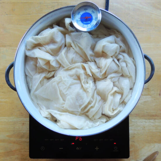 Overhead view of a large white pot on a black hotplate on a light wood table. Inside the pot is muslin fabric and water. A candy thermometer is attached to the pot.